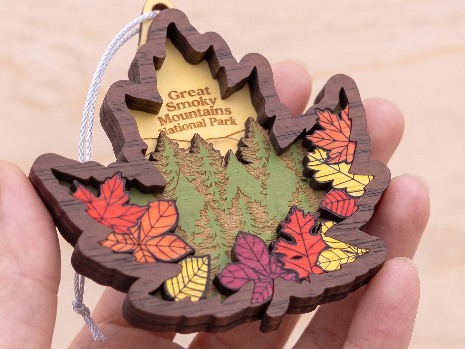 A hand holding a laser-engraved wood ornament for Great Smoky Mountains National Park, shaped like an autumn leaf
